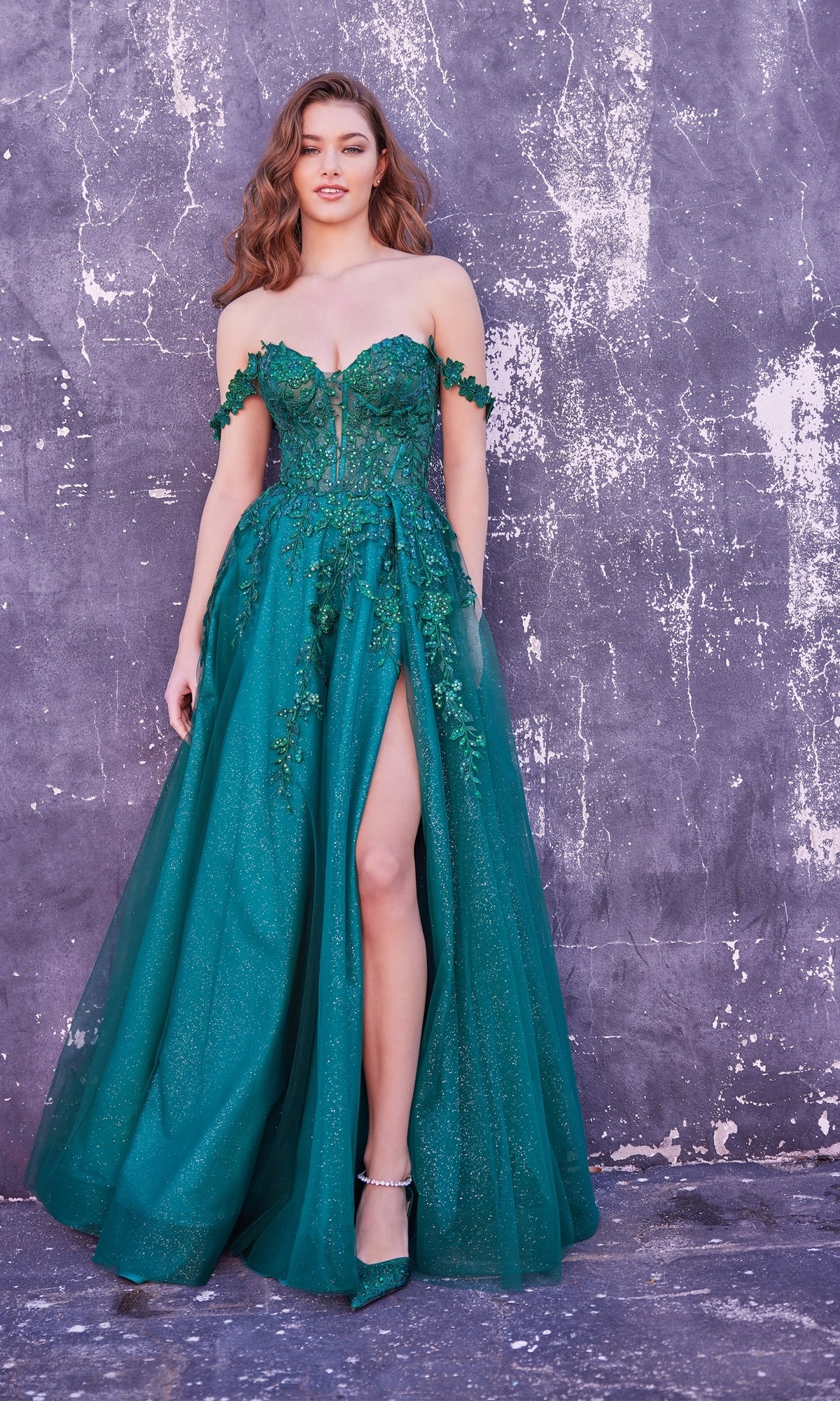 A woman stands against a textured gray wall, wearing the Ellie Wilde Off-Shoulder Long Prom Dress EW35101 in emerald green, featuring floral embellishments and a thigh-high slit.